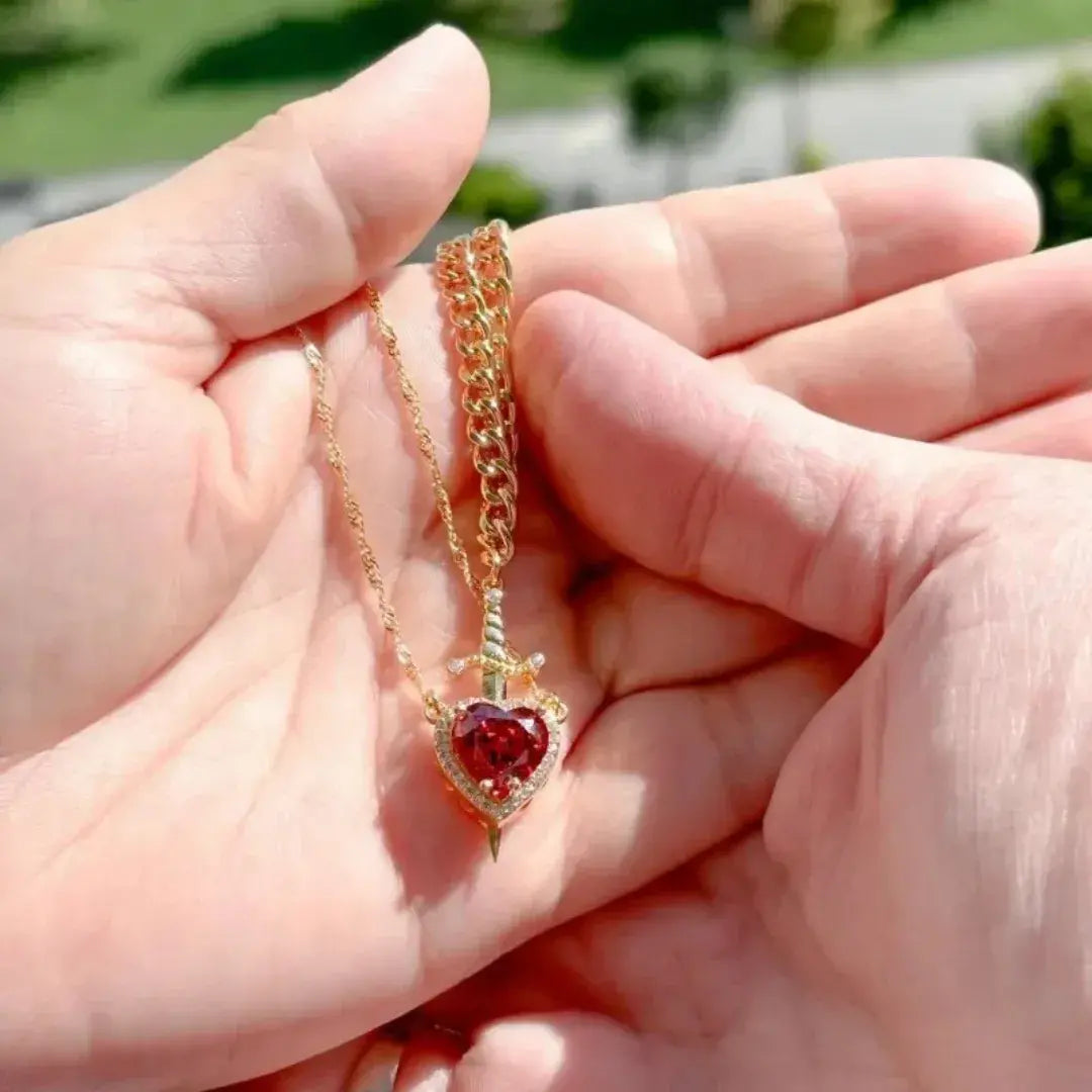 Colar dourado com pingente de coração vermelho sendo segurado na mão, brilho à luz natural – Romantizze