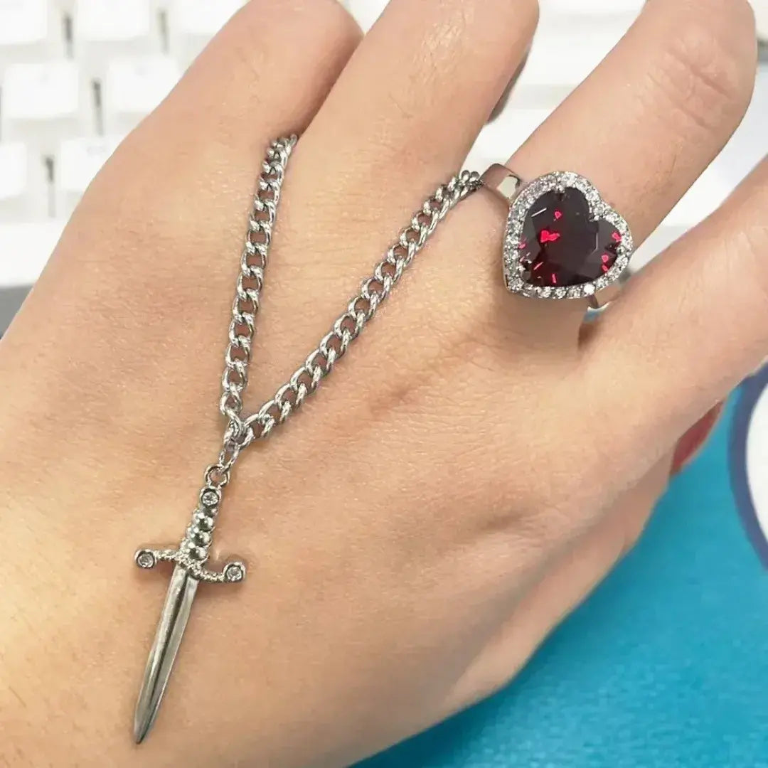 Colar prateado com pingente de espada ao lado de anel com pedra vermelha em forma de coração – Romantizze