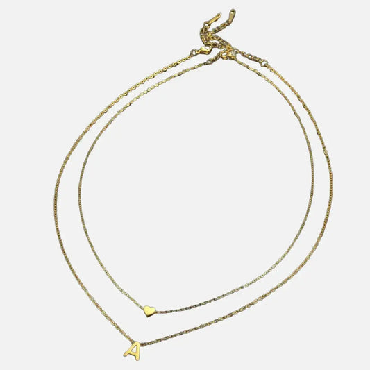 Colar com letra inicial e pingente de coração em aço inoxidável nas cores dourado e prata da Romantizze, sobre fundo branco.